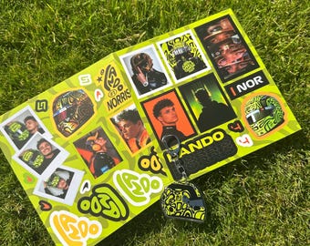 May include: A bright lime green rectangular sheet covered in various Lando Norris themed stickers, including photographs of the driver, racing helmets, and text graphics. A matching helmet-shaped keychain hangs over the bottom right corner.