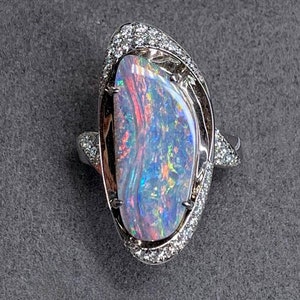May include: A silver-toned ring featuring a large, elongated, iridescent opal as the centerpiece. The opal displays vibrant flashes of red, blue, and green. The setting is surrounded by a halo of small, clear, sparkling stones set in metal.