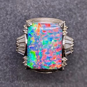 May include: A silver-toned ring featuring a large, rectangular, iridescent opal with vibrant flashes of red, blue, and green. The center stone is flanked by baguette-cut clear stones on each side, with small round clear stones accenting the corners.