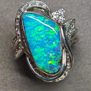 May include: A silver-toned ring featuring a large, irregular, teardrop-shaped opal with vibrant blue and green flashes. The opal is surrounded by a decorative metal setting encrusted with small, clear, round-cut stones along the band and top.