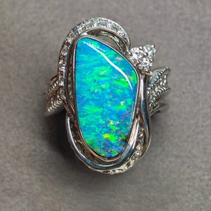 May include: A silver ring featuring a large, irregular, teardrop-shaped opal with vibrant blue and green flashes. The opal is surrounded by a decorative silver setting accented with small, clear, round-cut stones along the band and frame.