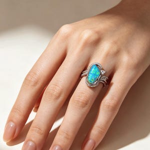 May include: A silver ring featuring a large, marquise-shaped iridescent blue and green opal as the centrepiece. The band is crafted with intricate metalwork and small, clear pav&eacute;-set stones that frame the central opal, creating a detailed design.