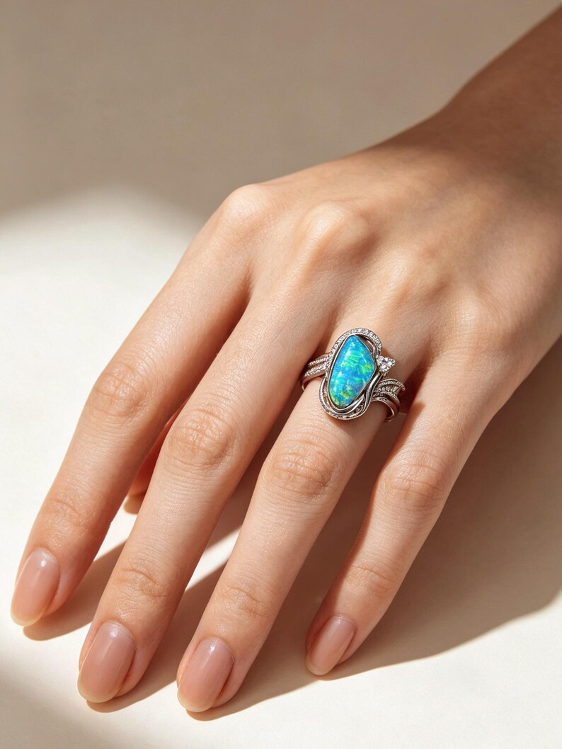 May include: A silver ring featuring a large, marquise-shaped iridescent blue and green opal as the centrepiece. The band is crafted with intricate metalwork and small, clear pav&eacute;-set stones that frame the central opal, creating a detailed design.