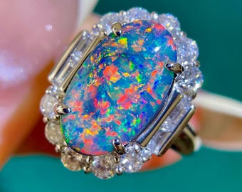 May include: A close-up of a ring featuring a large, oval-shaped opal with vibrant colors of blue, red, and green. The opal is surrounded by a halo of small, clear gemstones and baguette-cut stones, set in a gold band.