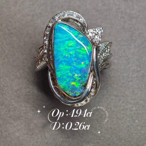 May include: A silver ring featuring a large, irregular, iridescent blue and green opal centre stone surrounded by small clear accent stones. The text Op: 4.94ct and D: 0.26ct is printed below the ring, indicating the weight of the stones.
