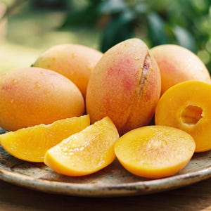 May include: A collection of whole and sliced yellow-orange mangoes arranged on a round, speckled ceramic plate. The fruit features smooth, thin skins with subtle red blushing, while the interior flesh is a vibrant, solid golden-yellow color.