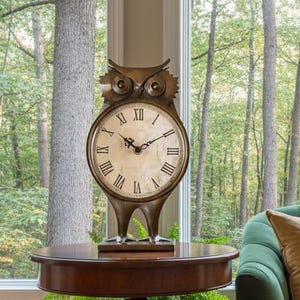 Può includere: Un orologio a forma di gufo con numeri romani è posto su un tavolo in legno scuro, accanto a un divano verde con cuscini dorati e fantasia. Un tappeto fantasia e una felce in vaso completano l'arredamento. Grandi finestre ad arco si affacciano su una foresta.