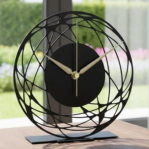 Puede incluir: Reloj de escritorio negro y dorado con un diseño esférico único. El reloj presenta una esfera negra con manecillas doradas y un marco decorativo de metal negro. Se apoya sobre una base rectangular negra, sobre una superficie de madera.