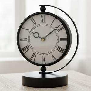 May include: A black and white analog clock with Roman numerals, placed on a small, light-coloured wooden table. The clock has a circular face and a curved metal frame. The hands indicate approximately 10:10. A decorative home decor item.