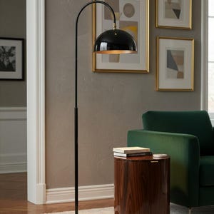 A burgundy floor lamp with a curved arm and a dome-shaped shade. The lamp is in a living room with a cream-colored sofa, gold pillows, and a wooden coffee table. The room has cream-colored curtains and a patterned wallpaper.