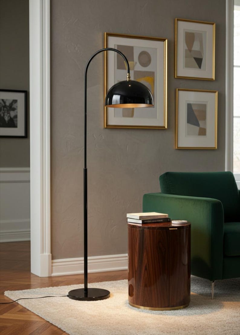 A burgundy floor lamp with a curved arm and a dome-shaped shade. The lamp is in a living room with a cream-colored sofa, gold pillows, and a wooden coffee table. The room has cream-colored curtains and a patterned wallpaper.