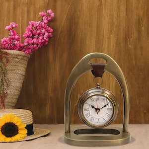 May include: A brass-coloured clock with a white face and black Roman numerals. The clock is suspended within a brass arch and has a brown leather strap. A straw hat with a sunflower and a basket of pink flowers are in the background.