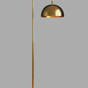 A gold floor lamp with a curved arm and a dome-shaped shade. The lamp is in a living room with a cream-colored sofa, gold pillows, and a wooden coffee table. The room has cream-colored curtains and a patterned wallpaper.