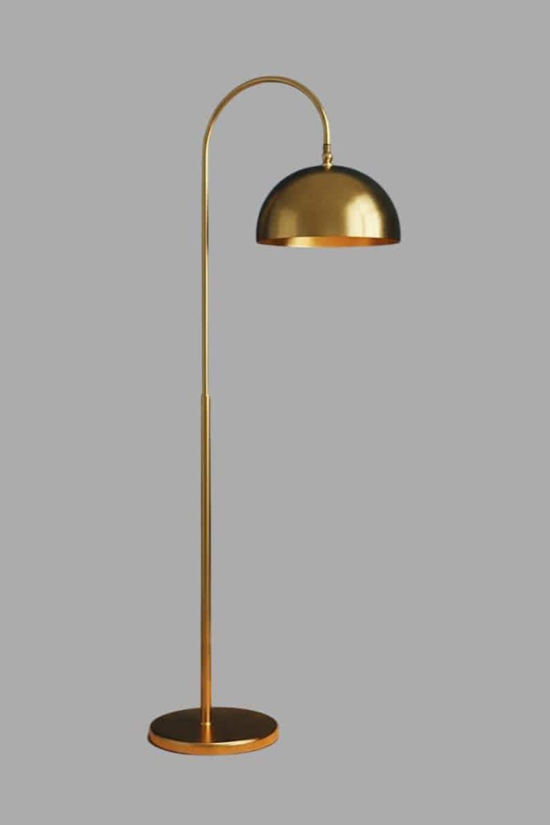A gold floor lamp with a curved arm and a dome-shaped shade. The lamp is in a living room with a cream-colored sofa, gold pillows, and a wooden coffee table. The room has cream-colored curtains and a patterned wallpaper.
