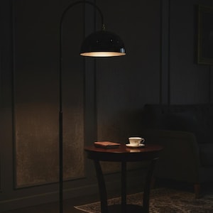 A burgundy floor lamp with a curved arm and a dome-shaped shade. The lamp is in a living room with a cream-colored sofa, gold pillows, and a wooden coffee table. The room has cream-colored curtains and a patterned wallpaper.