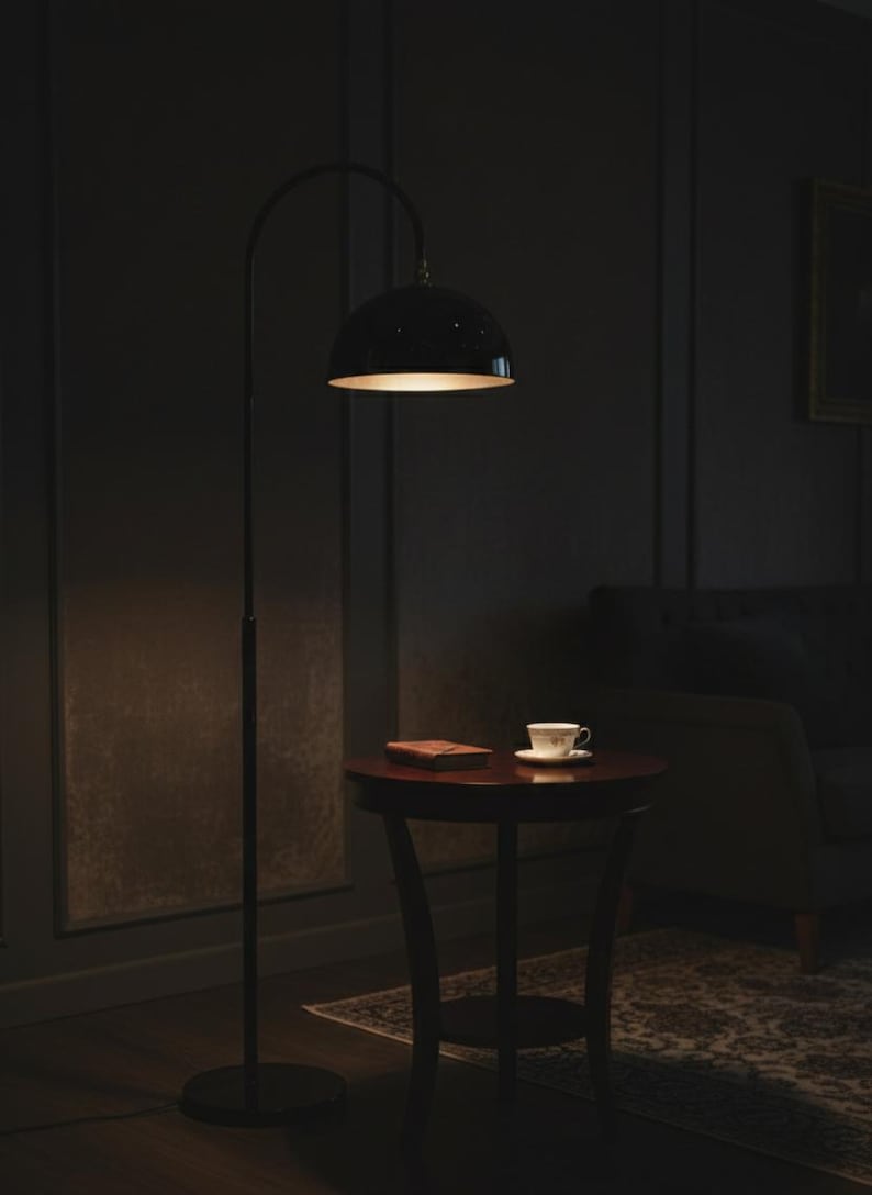 A burgundy floor lamp with a curved arm and a dome-shaped shade. The lamp is in a living room with a cream-colored sofa, gold pillows, and a wooden coffee table. The room has cream-colored curtains and a patterned wallpaper.