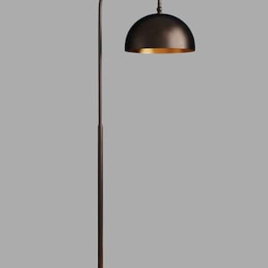 A burgundy floor lamp with a curved arm and a dome-shaped shade. The lamp is in a living room with a cream-colored sofa, gold pillows, and a wooden coffee table. The room has cream-colored curtains and a patterned wallpaper.