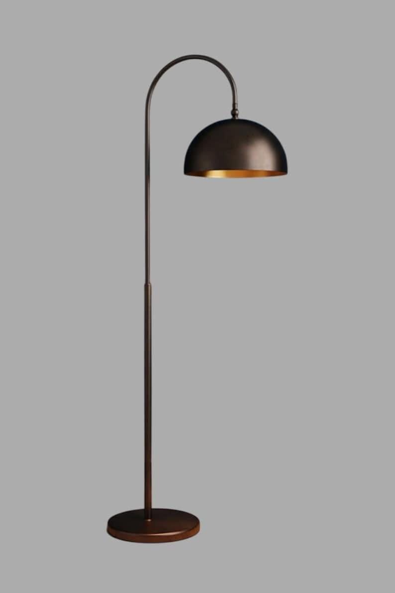 A burgundy floor lamp with a curved arm and a dome-shaped shade. The lamp is in a living room with a cream-colored sofa, gold pillows, and a wooden coffee table. The room has cream-colored curtains and a patterned wallpaper.
