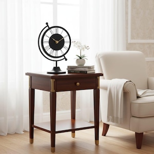A dark brown wooden side table with a drawer and a lower shelf. A black globe-shaped clock with Roman numerals sits on top, along with a small white potted plant and books. The table has brass accents and stands 24 inches tall.