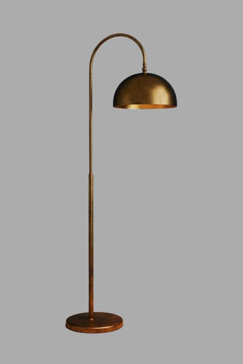 A antique floor lamp with a curved arm and a dome-shaped shade. The lamp is in a living room with a cream-colored sofa, gold pillows, and a wooden coffee table. The room has cream-colored curtains and a patterned wallpaper.