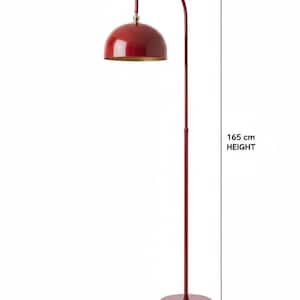 A burgundy floor lamp with a curved arm and a dome-shaped shade. The lamp is in a living room with a cream-colored sofa, gold pillows, and a wooden coffee table. The room has cream-colored curtains and a patterned wallpaper.