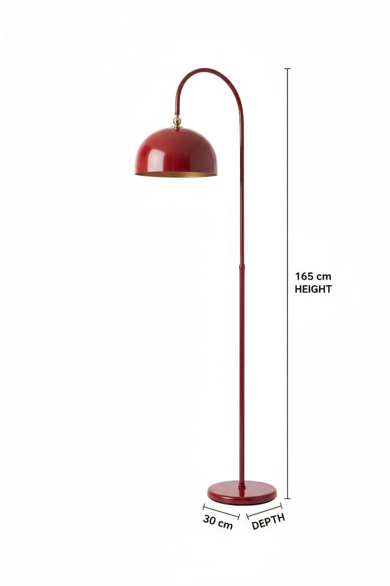 A burgundy floor lamp with a curved arm and a dome-shaped shade. The lamp is in a living room with a cream-colored sofa, gold pillows, and a wooden coffee table. The room has cream-colored curtains and a patterned wallpaper.