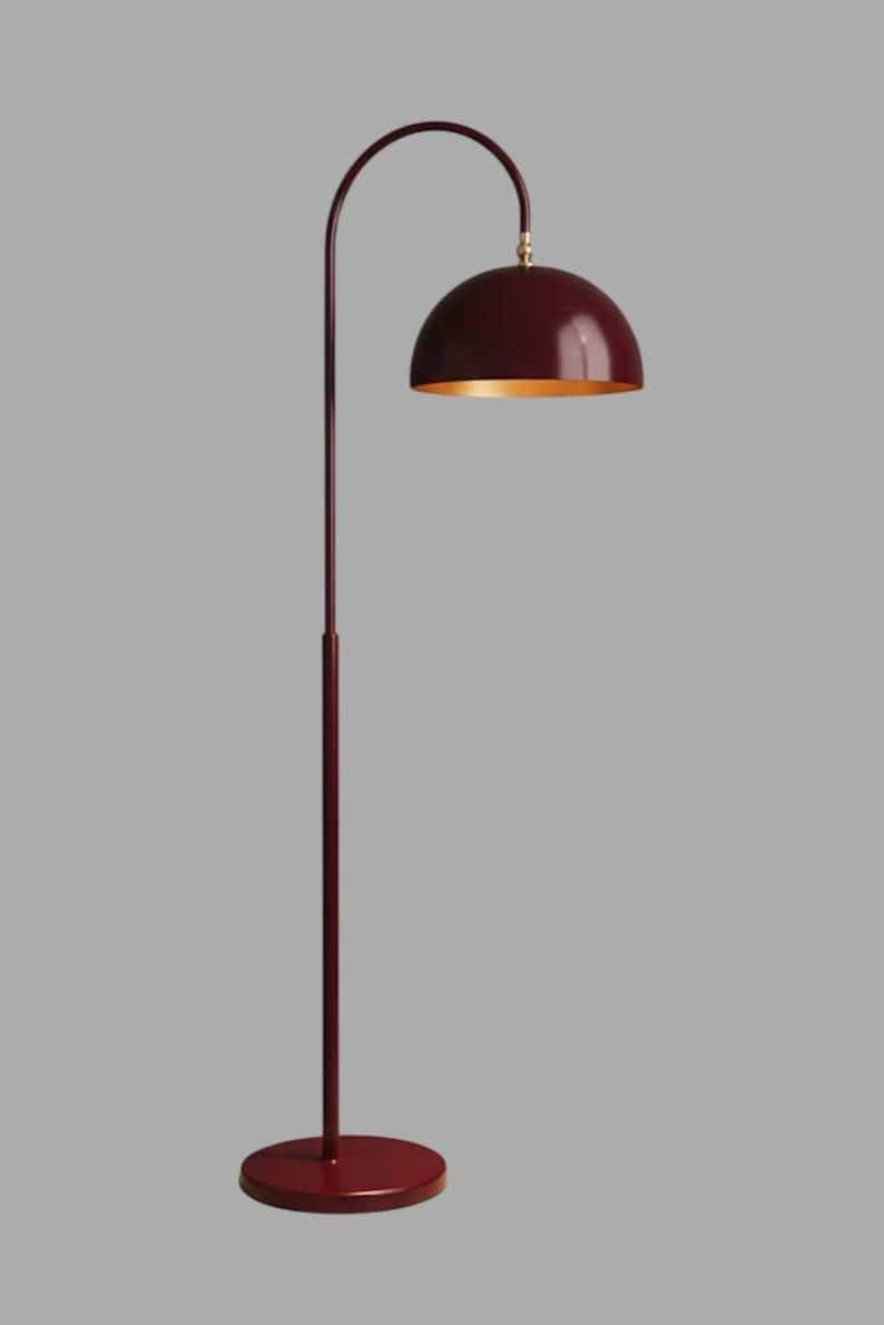 A burgundy floor lamp with a curved arm and a dome-shaped shade. The lamp is in a living room with a cream-colored sofa, gold pillows, and a wooden coffee table. The room has cream-colored curtains and a patterned wallpaper.