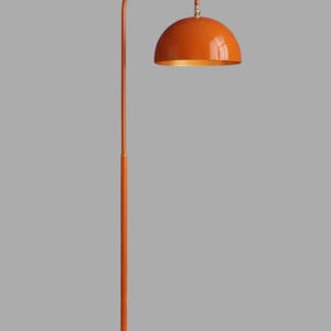 A orange floor lamp with a curved arm and a dome-shaped shade. The lamp is in a living room with a cream-colored sofa, gold pillows, and a wooden coffee table. The room has cream-colored curtains and a patterned wallpaper.