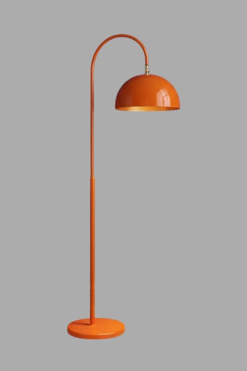 A orange floor lamp with a curved arm and a dome-shaped shade. The lamp is in a living room with a cream-colored sofa, gold pillows, and a wooden coffee table. The room has cream-colored curtains and a patterned wallpaper.
