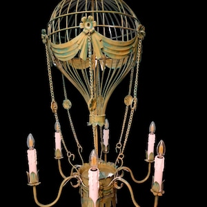 Może przedstawiać: Żyrandol w stylu antycznym w kształcie balonu na ogrzane powietrze. Metalowa rama pomalowana jest na złoto z turkusowymi akcentami. Posiada sześć świateł w kształcie świec z różowymi żarówkami i ozdobnymi łańcuchami.