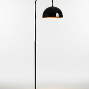 A gloss black floor lamp with a curved arm and a dome-shaped shade. The lamp is in a living room with a cream-colored sofa, gold pillows, and a wooden coffee table. The room has cream-colored curtains and a patterned wallpaper.
