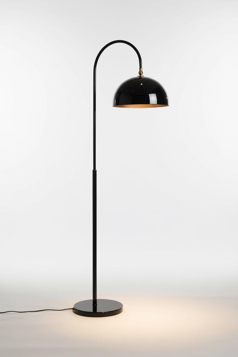 A gloss black floor lamp with a curved arm and a dome-shaped shade. The lamp is in a living room with a cream-colored sofa, gold pillows, and a wooden coffee table. The room has cream-colored curtains and a patterned wallpaper.