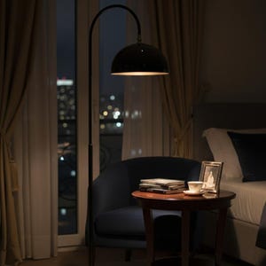 A burgundy floor lamp with a curved arm and a dome-shaped shade. The lamp is in a living room with a cream-colored sofa, gold pillows, and a wooden coffee table. The room has cream-colored curtains and a patterned wallpaper.