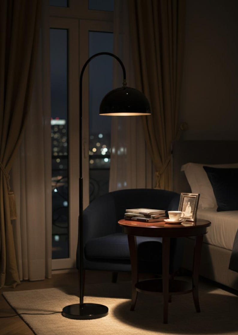 A burgundy floor lamp with a curved arm and a dome-shaped shade. The lamp is in a living room with a cream-colored sofa, gold pillows, and a wooden coffee table. The room has cream-colored curtains and a patterned wallpaper.
