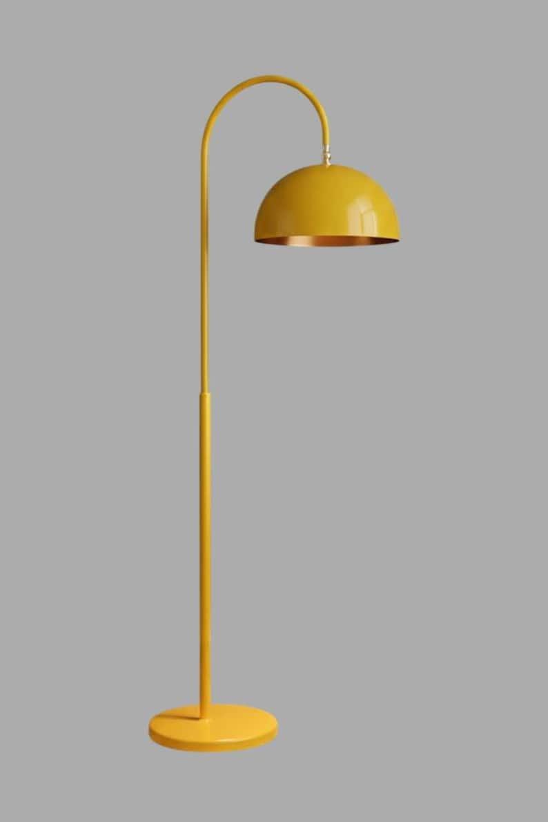A yellow floor lamp with a curved arm and a dome-shaped shade. The lamp is in a living room with a cream-colored sofa, gold pillows, and a wooden coffee table. The room has cream-colored curtains and a patterned wallpaper.