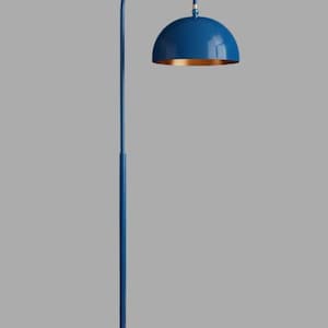 A blue floor lamp with a curved arm and a dome-shaped shade. The lamp is in a living room with a cream-colored sofa, gold pillows, and a wooden coffee table. The room has cream-colored curtains and a patterned wallpaper.