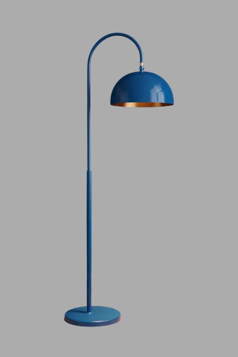 A blue floor lamp with a curved arm and a dome-shaped shade. The lamp is in a living room with a cream-colored sofa, gold pillows, and a wooden coffee table. The room has cream-colored curtains and a patterned wallpaper.