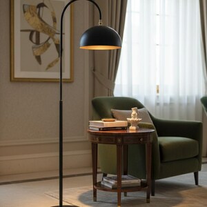 A black floor lamp with a curved arm and a dome-shaped shade. The lamp is in a living room with a cream-colored sofa, gold pillows, and a wooden coffee table. The room has cream-colored curtains and a patterned wallpaper.