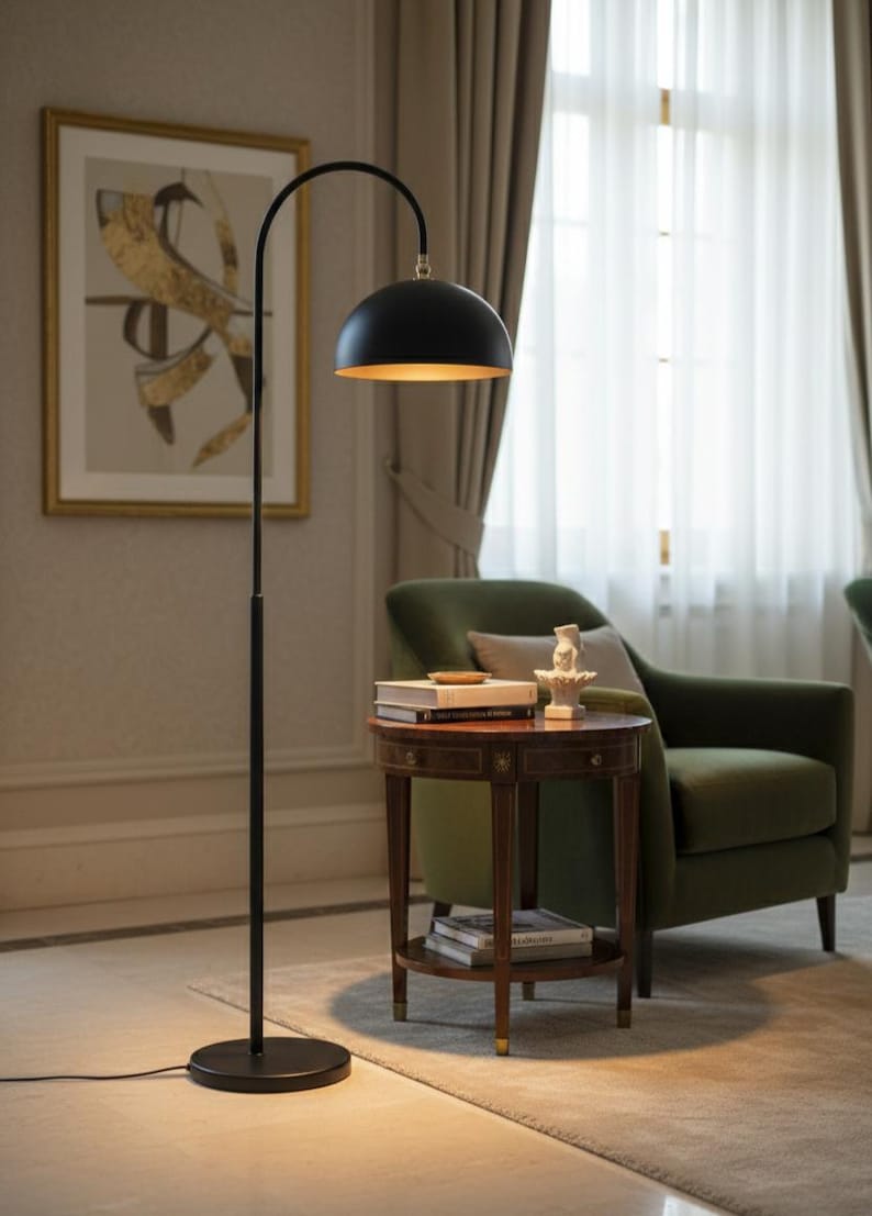 A black floor lamp with a curved arm and a dome-shaped shade. The lamp is in a living room with a cream-colored sofa, gold pillows, and a wooden coffee table. The room has cream-colored curtains and a patterned wallpaper.