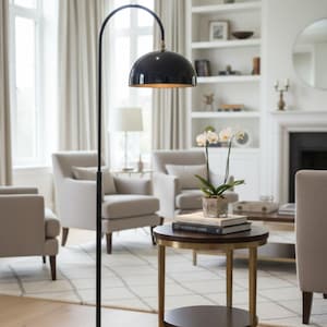 A burgundy floor lamp with a curved arm and a dome-shaped shade. The lamp is in a living room with a cream-colored sofa, gold pillows, and a wooden coffee table. The room has cream-colored curtains and a patterned wallpaper.