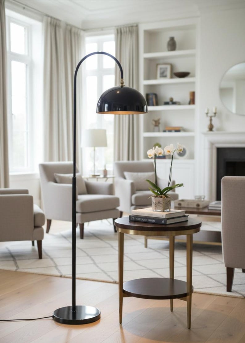A burgundy floor lamp with a curved arm and a dome-shaped shade. The lamp is in a living room with a cream-colored sofa, gold pillows, and a wooden coffee table. The room has cream-colored curtains and a patterned wallpaper.