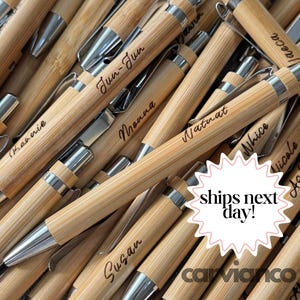 May include: A collection of bamboo pens with silver-toned metal clips and tips. Each pen features a unique name engraved in a cursive script on the barrel. The pens are arranged in a pile, showcasing the natural light wood grain texture.