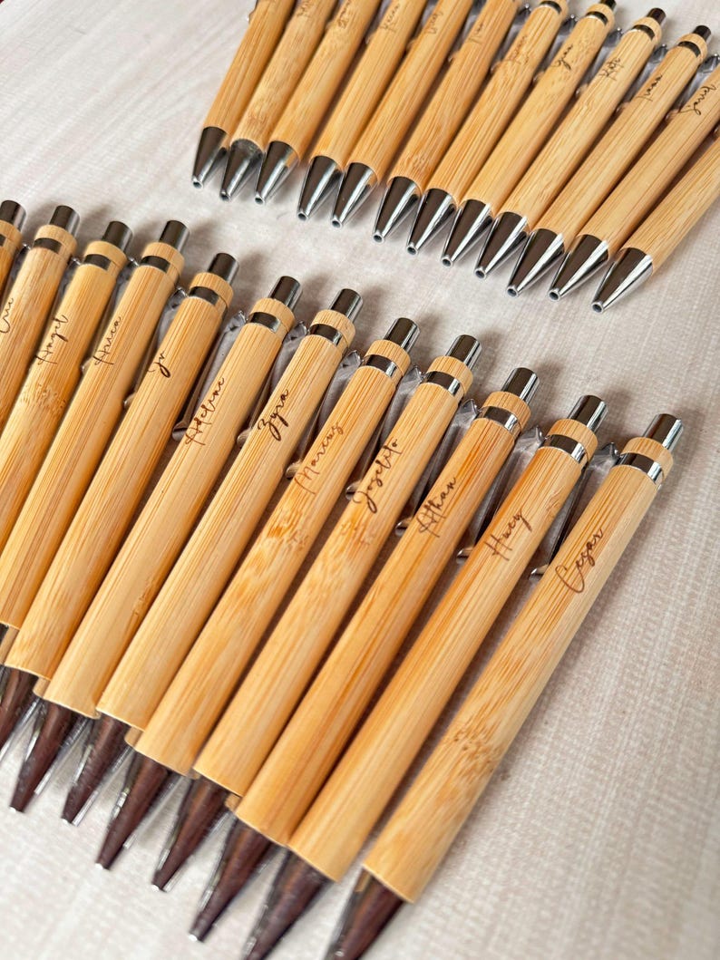 May include: A collection of bamboo ballpoint pens arranged in two rows. Each pen features a light brown natural wood grain barrel with a silver metal tip, clip, and click mechanism. Several pens are engraved with various names in cursive script.