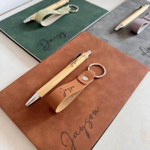 May include: A set consisting of a brown leather notebook, a bamboo pen, and a brown leather keychain, all personalized with the name Jayson in black script. The notebook cover, pen barrel, and keychain strap each feature the name Jayson printed.
