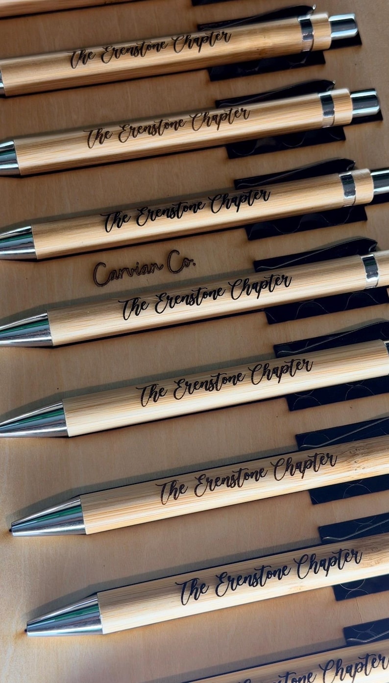 May include: A collection of light brown bamboo pens with silver-toned metal tips and clips. Each pen is engraved with the text The Erenstone Chapter in a black script font. One pen also features the engraved text Carvian Co. above the main title.