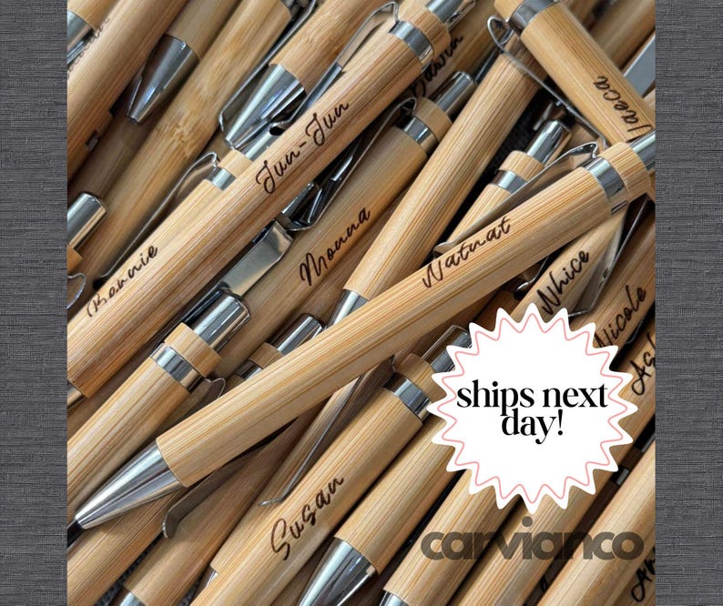 May include: A collection of bamboo pens with silver-toned metal clips and tips. Each pen features a unique name engraved in a cursive script on the barrel. The pens are arranged in a pile, showcasing the natural light wood grain texture.
