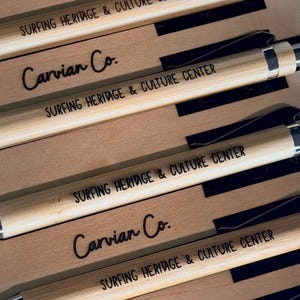 May include: A collection of bamboo pens arranged in rows, each featuring a silver-toned metal clip and tip. The pens are engraved with the text "SURFING HERITAGE & CULTURE CENTER" and are displayed alongside cardboard inserts labeled "Carvian Co."