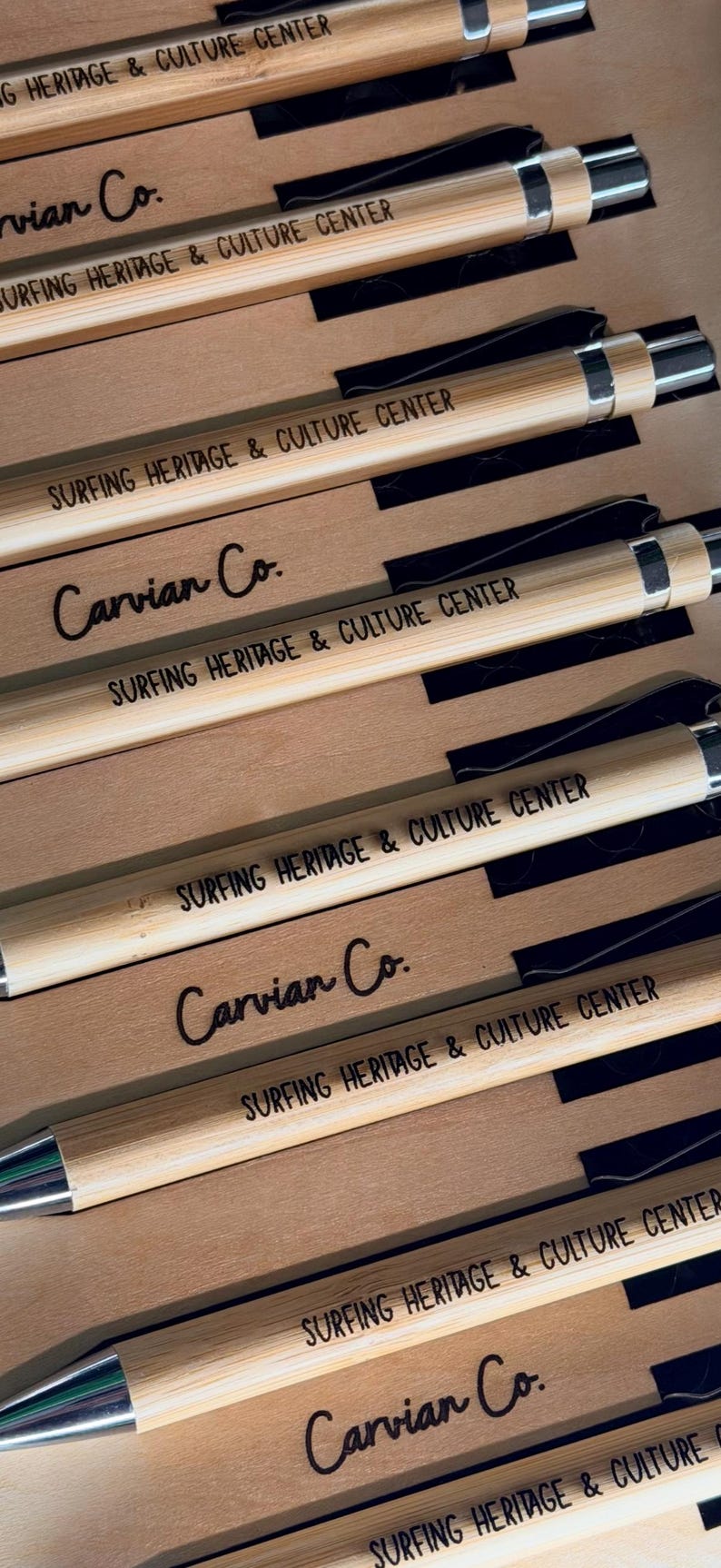 May include: A collection of bamboo pens arranged in rows, each featuring a silver-toned metal clip and tip. The pens are engraved with the text "SURFING HERITAGE & CULTURE CENTER" and are displayed alongside cardboard inserts labeled "Carvian Co."