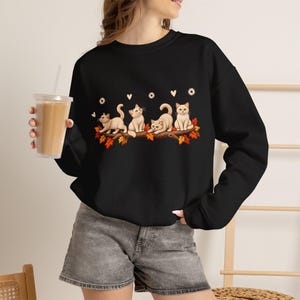 Può includere: Una felpa nera a maniche lunghe con una stampa grafica di quattro gattini dal pelo chiaro appollaiati su un ramo d'albero decorato con foglie autunnali arancioni e rosse, piccoli fiori bianchi e cuori, abbinata a pantaloncini di jeans grigi.