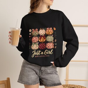 Può includere: Una felpa nera a maniche lunghe con una stampa grafica di nove zucche con vari motivi e fiocchi, accompagnata dal testo "Just a Girl WHO LOVES FALL" in un carattere chiaro in corsivo e stampatello sulla parte anteriore.